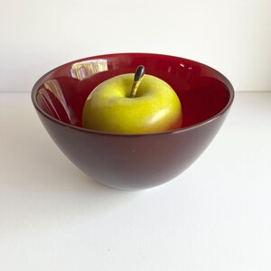 May include: A deep red bowl holds a green and yellow apple. The bowl has a smooth, glossy finish and a rounded shape. The apple has a long stem and a natural appearance. The bowl and apple are set against a white background.