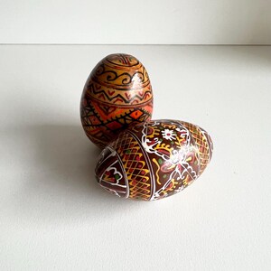 May include: Two decorated Easter eggs with detailed, colourful patterns. One egg displays orange, red, and brown designs, while the other has a brown base with white, yellow, and green accents. The eggs are likely hand-painted.