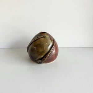 Peut inclure: Un petit vase en céramique fait à la main de forme arrondie. Le vase présente un mélange de couleurs, dont un bordeaux profond, un vert olive et un marron. Le design comprend un motif tourbillonnant texturé autour du vase.