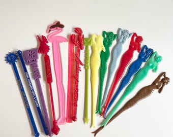 May include: A selection of colourful cocktail stirrers in various shapes, including mermaids, a flamingo, and a lobster. Some stirrers have text, such as "Dewar's" and "Aruba Caribbean Hotel & Casino". The stirrers are made of plastic.