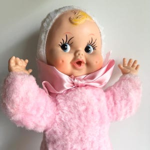 May include: A vintage baby doll with a soft pink plush body and a white bonnet. The doll has a painted face with blue eyes, rosy cheeks, and a pink satin bow. The doll's arms are raised, and it appears to be smiling.