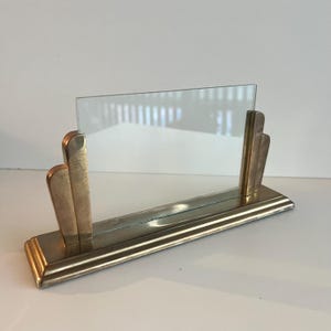 May include: A vintage Art Deco style picture frame. The frame is made of gold-toned metal with a rectangular base and two vertical supports holding a clear glass pane. The design is geometric and streamlined.