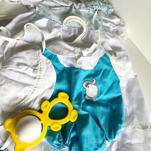 May include: A baby's outfit set including a white bonnet, a turquoise and white romper with an elephant applique, and a yellow rattle. The romper has a white collar and button closure. A white dress with floral accents hangs in the background.