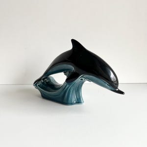 Könnte beinhalten: Eine schwarze und türkisfarbene Keramik-Delfinfigur, die aus dem Wasser springt. Der Delfin befindet sich in der Luft, mit ausgebreiteten Schwanz und Flossen. Die Basis der Figur ist eine Welle mit türkisfarbenem Überzug.