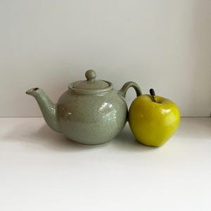 May include: A speckled, light green ceramic teapot with a curved spout, handle, and lid, next to a bright yellow apple. The teapot has a rounded body and a small knob on the lid. The apple has a dark stem.