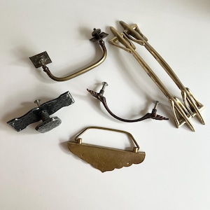 Peut inclure: Une collection de quincaillerie de meubles vintage, comprenant des poignées et des tirettes en laiton et en métal foncé. L'assortiment présente diverses formes et designs, tels que des poignées courbes, des tirettes en forme de flèche et une tirette rectangulaire décorative.