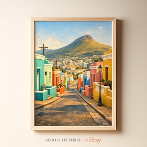 Può includere: Una stampa artistica incorniciata raffigura una vivace scena di strada con edifici nei toni del turchese, arancione, rosa e giallo. Una montagna incombe sullo sfondo sotto un cielo blu. La stampa è in una cornice di legno chiaro. Il testo in basso recita "SKYWARD ART PRINTS | ON Etsy."