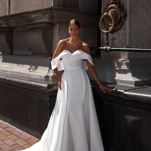 Op de afbeelding: Een witte, off-the-shoulder trouwjurk met een zwierige rok en sleep. De jurk heeft een getailleerd lijfje en een eenvoudig, elegant ontwerp. Het model draagt een delicate ketting. De achtergrond is een stenen gebouw.