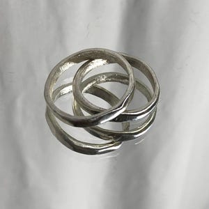 Könnte beinhalten: Drei ineinandergreifende silberne Ringe mit einem strukturierten, organischen Design. Die Ringe sind ineinander verschlungen und erzeugen einen einzigartigen und modernen Look. Die Ringe werden auf einer reflektierenden Oberfläche präsentiert.