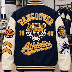May include: A navy blue varsity jacket with cream-colored sleeves. The back features a large tiger head graphic, the text "VANCOUVER" and "Athletics," and the numbers "19" and "48." The sleeves have patches including "VAN CHAMPS" and "VARSITY ATHLETICS."