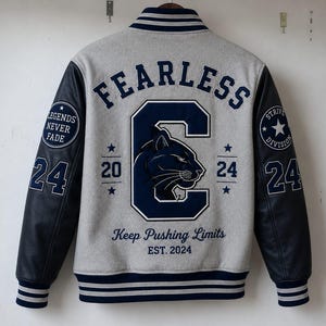 Op de afbeelding: Een grijs varsity-jack met donkerblauwe mouwen, voorzien van de tekst FEARLESS, een grote letter C met een panterafbeelding, de cijfers 20 en 24, de zin Keep Pushing Limits, EST. 2024, en ronde patches met de teksten Legends Never Fade en Strive Division.