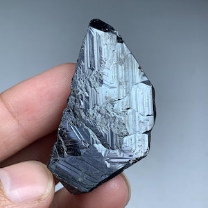 May include: A dark, irregularly shaped mineral specimen with a reflective, metallic luster. The surface displays linear striations and a rough texture, with a deep black color. The mineral is held in a hand against a neutral background.