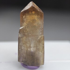 May include: A translucent, amber-colored crystal with a pointed top. The crystal has a rectangular body with a few small protrusions. It sits on a small, rounded, purple base. The crystal is set against a white surface and a dark gray background.