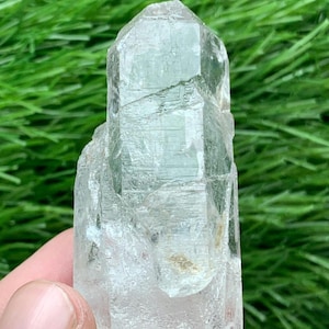 Puede incluir: Un cristal transparente de color verde claro con una parte superior facetada y un cuerpo texturizado. El cristal se sostiene sobre un fondo de hierba verde. La superficie del cristal tiene imperfecciones naturales.