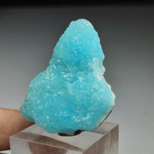 Puede incluir: Un vibrante espécimen mineral de color azul claro con una estructura cristalina. El mineral está montado sobre una base rectangular transparente. El color es un turquesa llamativo, con una superficie rugosa y texturizada. La forma general es irregular y orgánica.