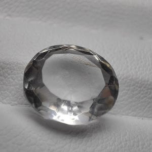 Puede incluir: Una gema transparente, tallada en forma ovalada y con múltiples facetas. La gema refleja la luz, creando un efecto brillante. La piedra está colocada sobre un fondo blanco.