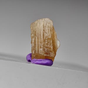 May include: A translucent, amber-colored crystal specimen, possibly a gemstone, is displayed. It is mounted on a small, rounded, purple base. The crystal has a rough, textured surface and a rectangular shape. The background is a soft gray.