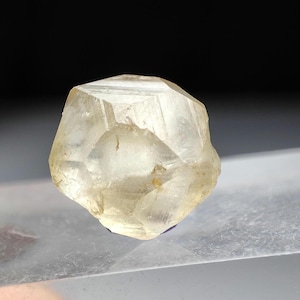 Pode incluir: Cristal facetado, translúcido, amarelo claro com forma geométrica. O cristal tem uma textura ligeiramente áspera e parece iluminado por dentro. O fundo é escuro, realçando a forma do cristal.