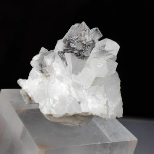 May include: A cluster of clear quartz crystals with a dark gray mineral inclusion. The quartz crystals are translucent and have sharp, pointed terminations. The mineral inclusion is a dark gray color and is located in the center of the quartz cluster. The quartz is mounted on a clear acrylic stand.