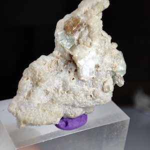 May include: A raw mineral specimen featuring a textured, off-white, and beige crystalline matrix. Embedded within the upper section are small, translucent, pale green crystals and a single, distinct, amber-colored crystal formation.