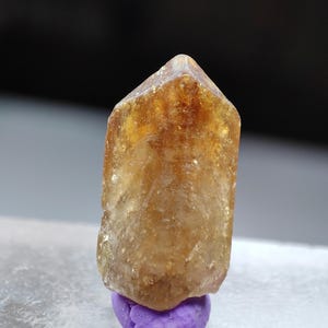 May include: A golden-brown crystal with a pointed top, resting on a small, purple base. The crystal has a rough, textured surface and a translucent appearance. The lighting highlights the crystal's natural color variations, creating a warm, earthy aesthetic.