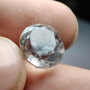 Puede incluir: Una gema clara y facetada con un interior ligeramente nublado. La piedra de forma ovalada se sostiene entre dos dedos, mostrando sus múltiples facetas y la refracción de la luz. La gema parece ser un cristal azul claro o incoloro.