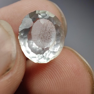 Puede incluir: Una gema facetada de forma ovalada con una apariencia pálida y translúcida. La gema tiene una forma ligeramente irregular y un sutil patrón interno. Las facetas capturan la luz, creando un efecto brillante. La gema se sostiene entre los dedos.