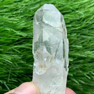 Puede incluir: Un cristal de cuarzo transparente con una punta. El cristal es translúcido con estructuras internas visibles y ligeras imperfecciones. El fondo es de un verde vibrante, creando un contraste con la claridad del cristal.
