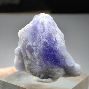 May include: A raw, unpolished tanzanite crystal specimen featuring a vibrant violet-blue hue. The mineral displays a jagged, crystalline structure with translucent sections and opaque white and grey inclusions at the base, set upon a clear block.