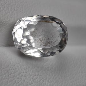 Puede incluir: Una gema clara, de forma ovalada y con múltiples facetas. La gema está tallada para reflejar la luz, creando un efecto brillante. La piedra se encuentra sobre un fondo neutro, resaltando su claridad y brillo. La gema parece ser un cristal de cuarzo.