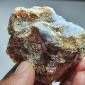 May include: A close-up of a rough, natural stone with a mix of colors. The stone features areas of light blue, beige, and brown, with a textured surface. The stone appears to be a mineral specimen.