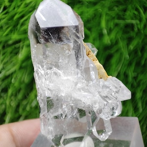 Puede incluir: Un grupo de cristales de cuarzo transparente con un interior ahumado de color gris oscuro. El cristal está montado sobre una base rectangular transparente, con un fondo verde. El cuarzo tiene múltiples facetas y un aspecto natural y en bruto.