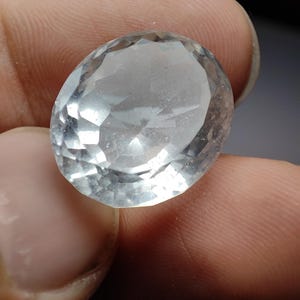 Puede incluir: Una gema clara, de corte ovalado, con múltiples facetas. La gema se sostiene entre los dedos, mostrando su claridad y el juego de la luz en su superficie. La piedra parece ser de un color claro y translúcido.