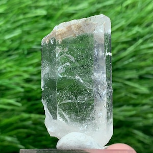 Puede incluir: Un cristal transparente y rectangular con una parte superior rugosa y texturizada y un cuerpo liso y pulido. El cristal está montado sobre una pequeña base gris, con un fondo de hierba verde borrosa.