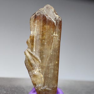 May include: A translucent, amber-colored crystal with a rough, textured surface. The crystal is mounted on a small, round, purple base. The crystal has a pointed top and a slightly irregular shape, with visible striations.