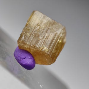 May include: A close-up of a rough, golden-brown mineral specimen with visible crystalline structures. A small, smooth, oval-shaped purple stone rests beneath the larger mineral. The background is a soft, blurred gray.