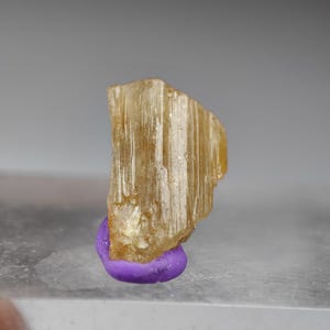 May include: A translucent, golden-yellow crystal specimen with a striated texture. The crystal is mounted on a small, rounded, purple base. The background is a neutral gray.