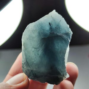 Puede incluir: Una gema de aguamarina en bruto, de color azul pálido. La piedra tiene una forma natural e irregular con inclusiones visibles. La gema se sostiene en una mano, con una fuente de luz brillante en el fondo. La gema es translúcida.