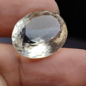 Puede incluir: Una gema facetada de forma ovalada con un tono amarillo pálido. La gema se sostiene entre los dedos, mostrando sus superficies pulidas y su claridad interna. La piedra refleja la luz, resaltando su talla y color.