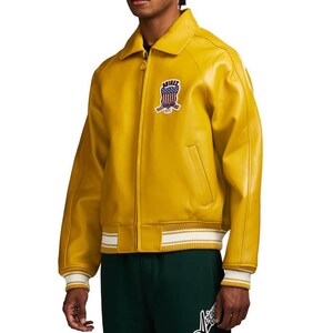 May include: Mustard yellow leather jacket with a zip-up front, a collar, and a patch on the chest. The jacket has white and yellow striped cuffs and a white and yellow striped hem. The jacket has a classic bomber jacket style.