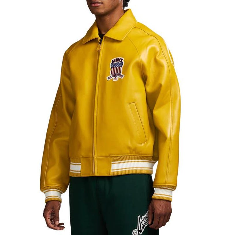 May include: Mustard yellow leather jacket with a zip-up front, a collar, and a patch on the chest. The jacket has white and yellow striped cuffs and a white and yellow striped hem. The jacket has a classic bomber jacket style.