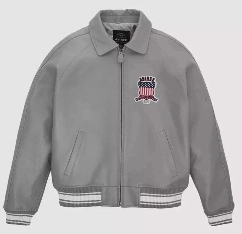 Puede incluir: Chaqueta bomber gris con cremallera, cuello y pu&ntilde;os y dobladillo acanalados. La chaqueta presenta un parche con detalles rojos, blancos y azules y la marca "AVIREX". La chaqueta tiene dos bolsillos delanteros.