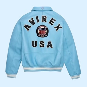 Puede incluir: Chaqueta de cuero azul claro con la palabra "AVIREX" arqueada sobre los hombros. Debajo, un parche con las palabras "ALL AMERICANS" y "USA". La chaqueta tiene pu&ntilde;os a rayas blancas y azules.