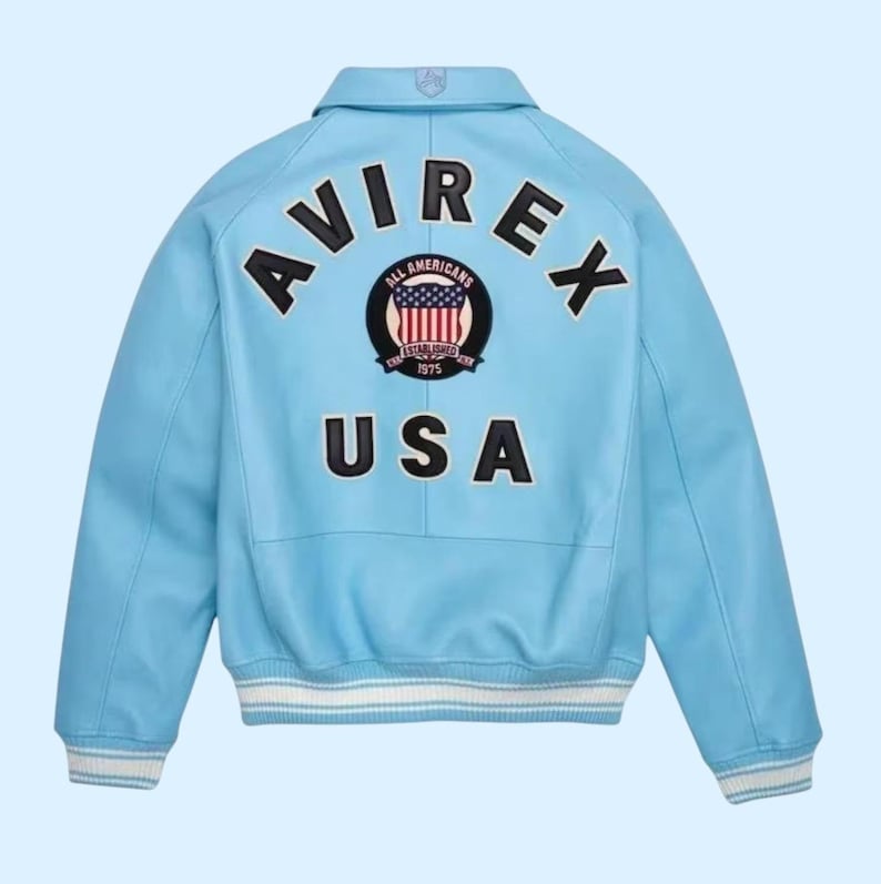Puede incluir: Chaqueta de cuero azul claro con la palabra "AVIREX" arqueada sobre los hombros. Debajo, un parche con las palabras "ALL AMERICANS" y "USA". La chaqueta tiene pu&ntilde;os a rayas blancas y azules.