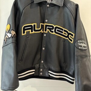 Puede incluir: Chaqueta de cuero negra con la palabra "AUREX" en letras amarillas y negras en el pecho. La chaqueta tiene cuello, pu&ntilde;os y dobladillo acanalados con rayas blancas. Un parche de dibujos animados est&aacute; en la manga izquierda y un parche "PALACE" en la manga derecha.