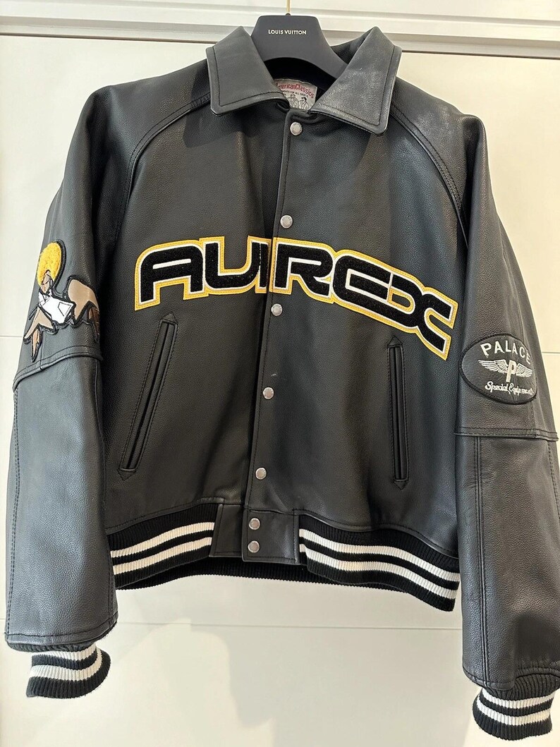 Puede incluir: Chaqueta de cuero negra con la palabra "AUREX" en letras amarillas y negras en el pecho. La chaqueta tiene cuello, pu&ntilde;os y dobladillo acanalados con rayas blancas. Un parche de dibujos animados est&aacute; en la manga izquierda y un parche "PALACE" en la manga derecha.