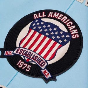 Puede incluir: Parche bordado circular con un dise&ntilde;o de la bandera estadounidense, las palabras "ALL AMERICANS" y "ESTABLISHED 1975 N.Y." en rojo y blanco. El parche tiene un borde negro y est&aacute; sobre un fondo azul claro.