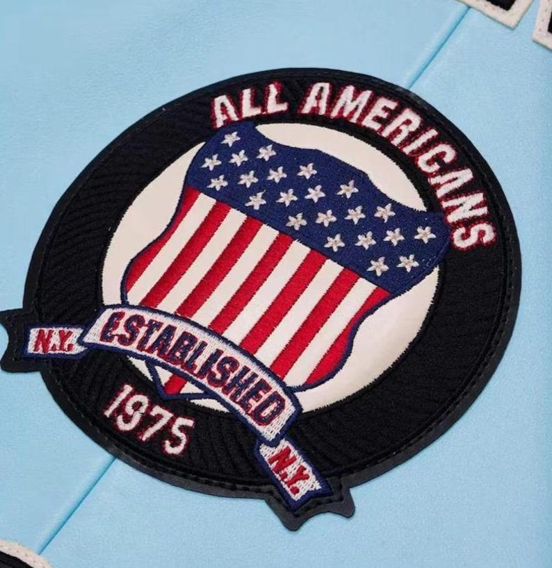 Puede incluir: Parche bordado circular con un dise&ntilde;o de la bandera estadounidense, las palabras "ALL AMERICANS" y "ESTABLISHED 1975 N.Y." en rojo y blanco. El parche tiene un borde negro y est&aacute; sobre un fondo azul claro.