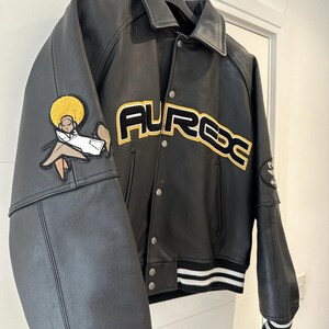 Puede incluir: Chaqueta universitaria de cuero negro con la palabra "AURDC" en letras amarillas y blancas en el pecho. La chaqueta tiene un dobladillo y pu&ntilde;os a rayas blancas y negras. Un parche de una persona est&aacute; en la manga izquierda.