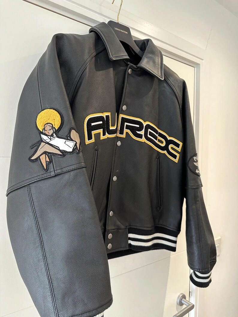 Puede incluir: Chaqueta universitaria de cuero negro con la palabra "AURDC" en letras amarillas y blancas en el pecho. La chaqueta tiene un dobladillo y pu&ntilde;os a rayas blancas y negras. Un parche de una persona est&aacute; en la manga izquierda.